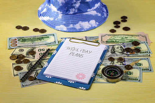 Financial plans for vacation concept. Blue hat, notebook with pencil, compass, hundred dollar bills and pennies on the table.