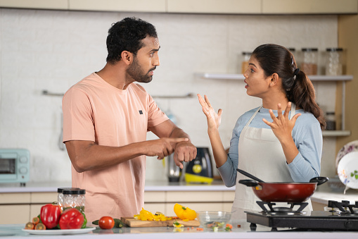 Indian Angry Husband arguing with wife while cooking at kitchen due to hungry - concept of family problems, relationship dispute and misunderstanding.