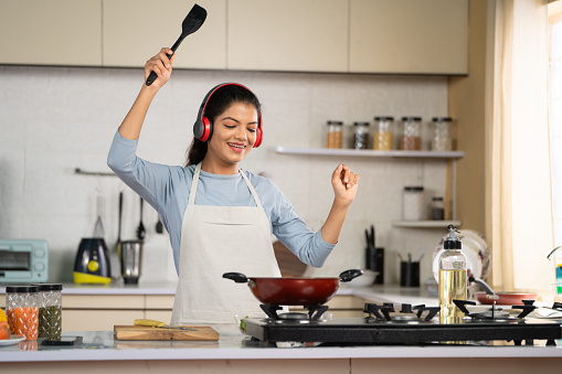 Joyful Dancing Indian woman cooking by listening songs on wireless headphones at kitchen - concept of weekend fun, food preparation and entertainment