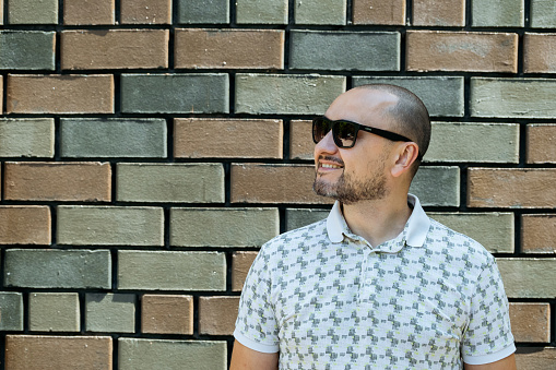 Cheerful adult male in sunglasses enjoys the sun against a textured wall.