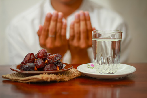Fast breaking meal or iftar dish with muslim man hands praying to Allah. Dates with a glass of mineral water on the table.Traditional Ramadan, fast breaking meal. Ramadan kareem fasting month concept.