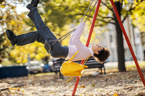 A cheerful trendy girl is swinging on a swing in city park.