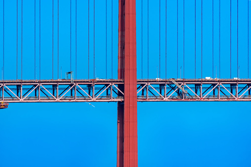 Frontal detail of part of the 25 de Abril suspension bridge with red steel cables supporting the structure with cars and vans driving on it to access Lisbon, under a clear blue sky and a pigeon flying