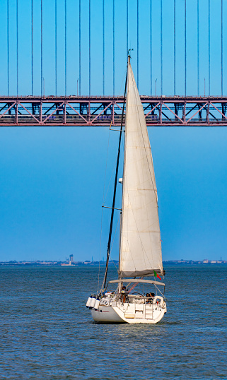 Tourist recreation sailboat sailing on the red steel 25 de Abril Suspension Bridge over the Tagus River in the city of Lisbon, Portugal, under a clear blue sky.