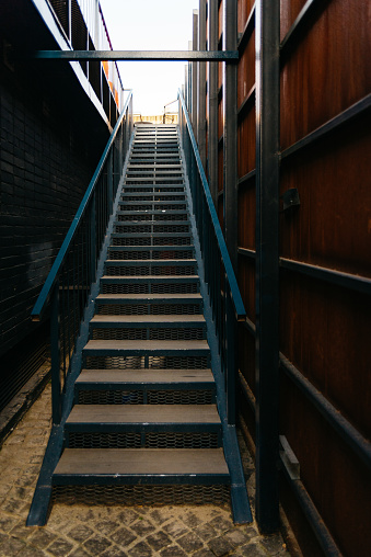 Looking up a metal staircase between dark walls, an urban escape leading to bright daylight above.