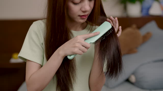Beautiful Asian woman sitting on bed inside bedroom, combing hair using hands pick up section hair, using comb comb hair make it flow beautifully, It's about styling hair so it doesn't get frizzy.