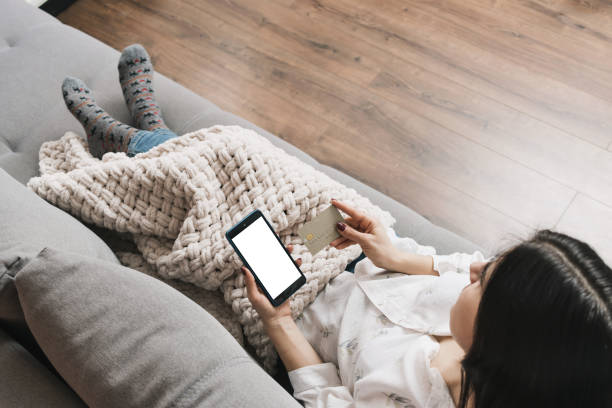 istock Young woman covered blanket, lying on a couch, holding credit card and using mobile phone with white blank screen, smartphone for online shopping or internet payment, top view 2007299414