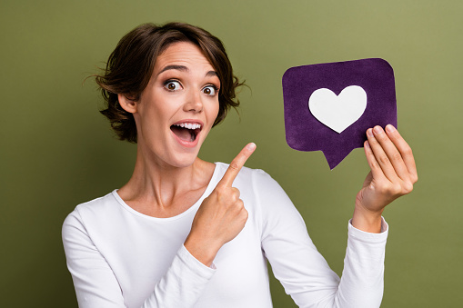 Photo portrait of lovely young lady hold point excited paper heart icon wear trendy white garment isolated on khaki color background.