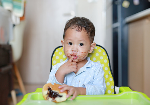 Happy Asian little baby boy sitting on children chair indoor eating bread with Stuffed Chocolate-filled dessert and Stained around her mouth.