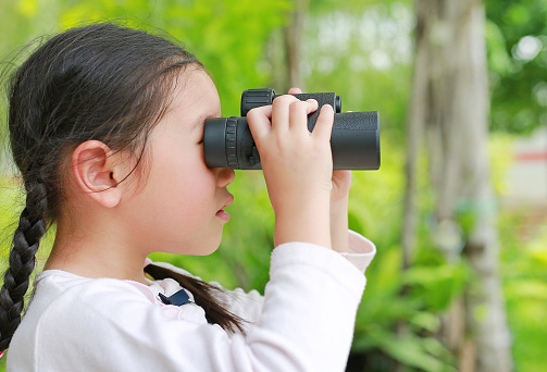 Little child girl in a field looking through binoculars in nature outdoor. Explore and adventure concept.