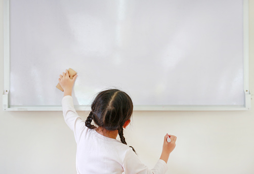 Little caucasian child girl using eraser on whiteboard in the classroom. White board with copy space for text. Rear view.