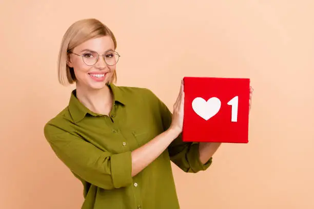 Photo of Photo of toothy beaming woman with bob hairstyle in glasses demonstarte one social media like in arms isolated on beige color background