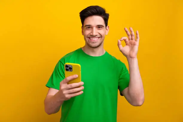 Photo of Photo of cheerful happy man wear green t shirt demonstrate nice feedback okey sign ad apple iphone 15 isolated on yellow color background