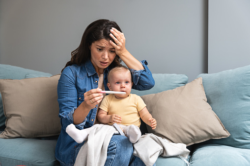 Young shocked mother holds a baby in her lap and a pregnancy test in her hand, she is surprised and in a panic because she is pregnant again, and she did not expect it. Woman issue concept