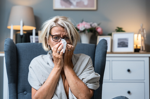 Older senior woman wrapped in a blanket wipes her nose with paper tissues, got sick because she didn't have money to pay for heating in her home or breakdown in the city heating network system.