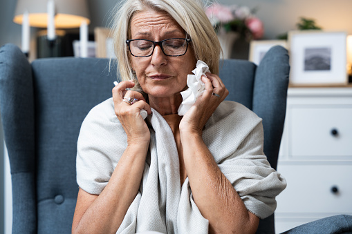 Older senior woman wrapped in a blanket wipes her nose with paper tissues, got sick because she didn't have money to pay for heating in her home or breakdown in the city heating network system.