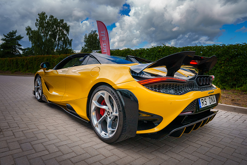 A vibrant yellow McLaren 765LT with a raised rear wing and sleek lines is parked outdoors, showcasing its aerodynamic design and performance features against a partly cloudy sky.