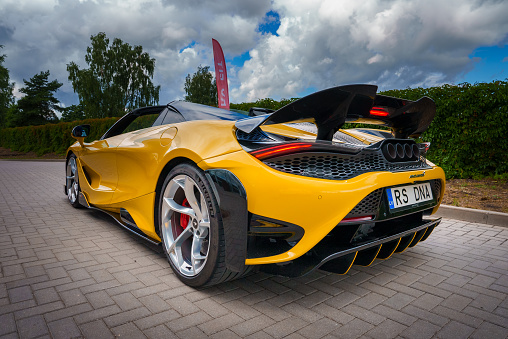 A bright yellow McLaren 765LT, with black details and silver rims, is angled to show its sleek design, active rear wing, and central exhaust. It's set outdoors with trees under a partly cloudy sky.
