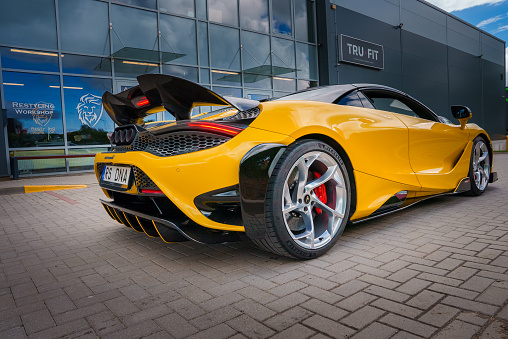 A vibrant yellow McLaren 765LT with a raised wing spoiler and hexagonal vents is parked outside a building, showcasing its aerodynamic design and sporty profile.
