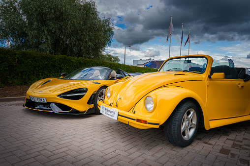 Yellow McLaren 765LT, aerodynamic, next to a classic VW Beetle convertible under clouds, shows car evolution. Flags indicate a windy day at a relaxed auto meet.