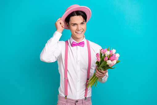 Photo of good mood pleasant guy wear white shirt hold tulips touching pink headwear on romantic date isolated on blue color background.