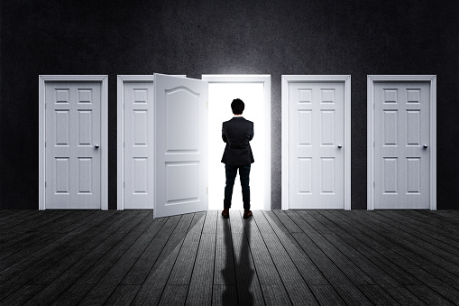 A person stands before an open doorway, surrounded by multiple closed doors, symbolizing decision-making and opportunity.