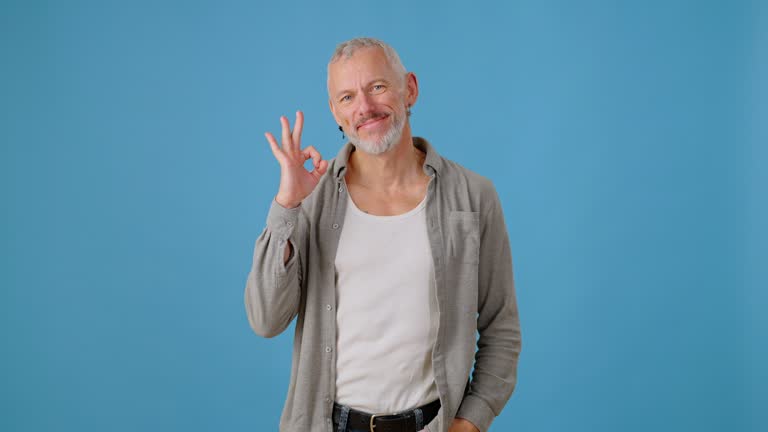 Mature man smiles gesturing with Okay sign against blue wall