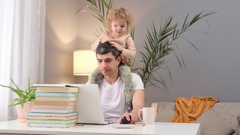 Baby and remote work. Business and parenthood. Child's influence on work. Confused Caucasian man working on laptop with baby daughter on his shoulders at home