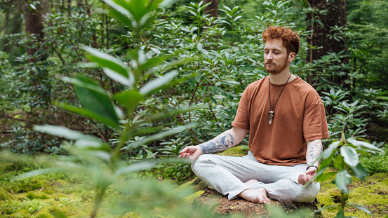 A medium close up side view of a young man who is sitting on a tree stump and meditation in a lush green forrest area in the North East of England. He is enjoying the ambient forrest noises as she enjoys the surroundings and feeling connected to nature.