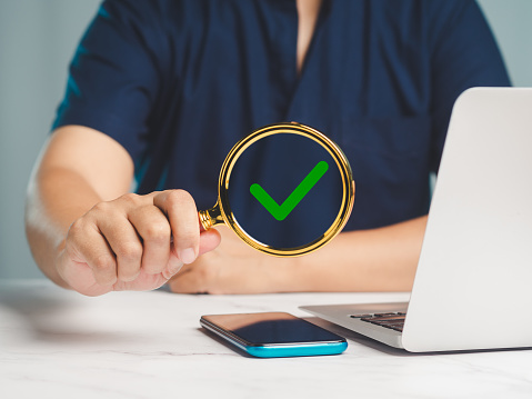 Businessman in a blue short-sleeved shirt holds a magnifying glass with a green check mark symbol while sitting at a desk in the office. Business and Approval project concept