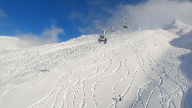 Two-Man Chairlift First-Person View Ascending on Sunny Blue Sky Day with Ski Tracks in Powder and Skiers Below