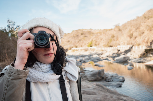 front view portrait of young latin photographer woman standing on the mountain looking through the viewfinder of her camera taking a photo, with the mountains in the background.