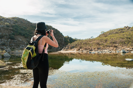 pretty young white hispanic latin tourist woman with backpack and cap standing in front of a lagoon taking pictures of two green mountains and the cloudy sky with her digital camera.