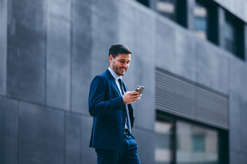 Portrait of a handsome man in a business suit, walking on the city street and looking down on his mobile phone. He is holding the smartphone, smiling and reading or writing a text message.