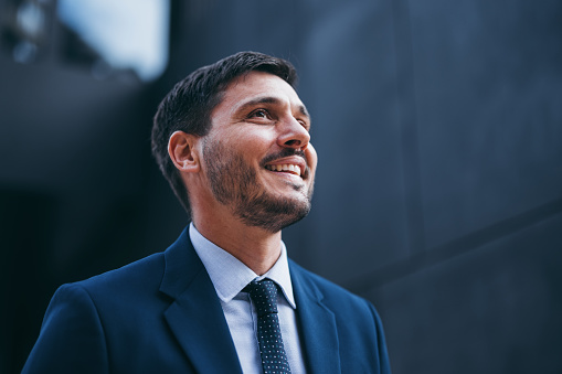 Close up shot of a handsome man in a business suit and tie, standing outdoors by the office building on the break from work. He is looking away and smiling.