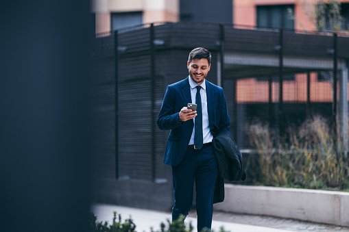 Handsome man in a suit and tie carrying a jacket, walking to his office or to a business meeting. He is holding mobile phone looking down, smiling and texting or reading a message.