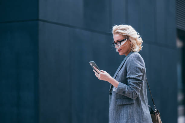 Beautiful Businesswoman Walking to Her Work Office and Reading a Message on Her Mobile Phone Beautiful blonde woman dressed formally for work or on her way to the meeting, walking on the city street between office buildings. She is reading a message on her mobile phone and smiling. older blonde woman stock pictures, royalty-free photos & images