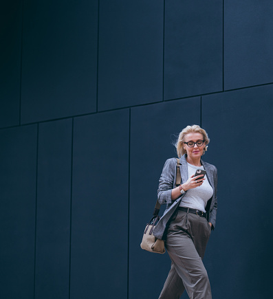Beautiful blonde woman dressed formally for work or on her way to the meeting, walking on the city street between office buildings. She is reading a message on her mobile phone and smiling.