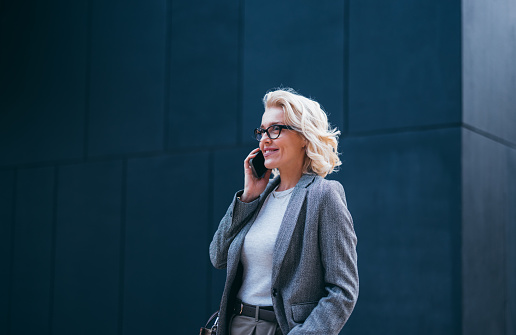 Close up shot of a beautiful blonde woman in business clothes and wearing glasses, standing by the office building and talking on her mobile phone. She is smiling and looking away.