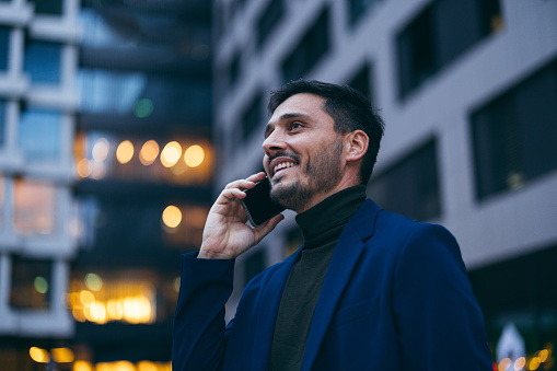 Close up shot of a handsome smiling businessman having a phone call outside his place of work. He is standing and talking while looking away.