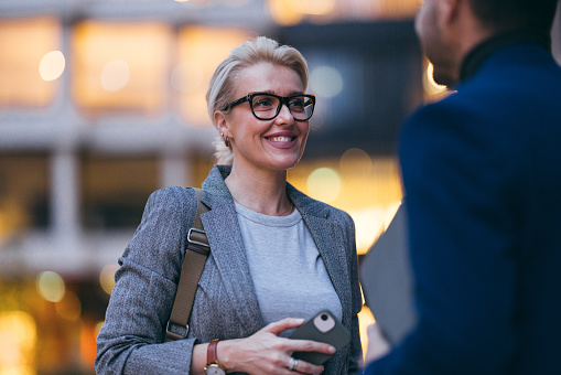 Close up of a beautiful blonde businesswoman wearing glasses. She is standing outdoors with an anonymous male colleague. She is smiling and looking at him.