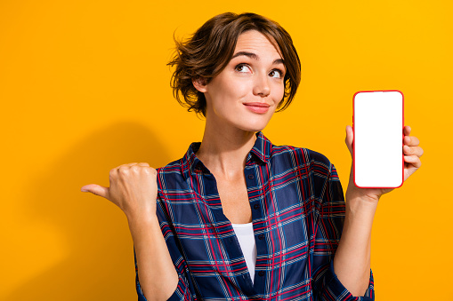 Portrait of attractive girl brunette hair show smartphone screen look directing at sale empty space isolated on yellow color background.