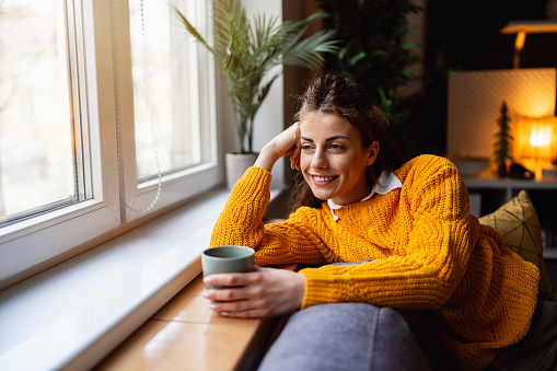 Young Caucasian woman relaxing on the sofa while drinking coffee and looking through window