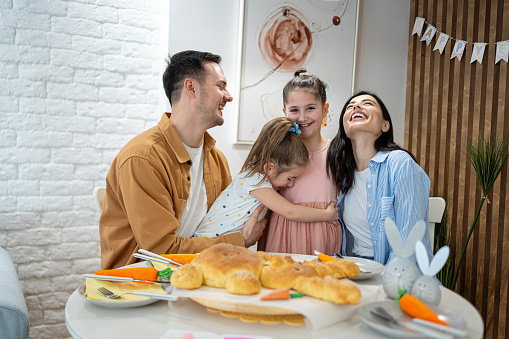 Young Caucasian family, parents with daughters celebrating an Easter, at their cozy and modern home, during an Easter lunch while having baked bread in the shape of the Easter Bunny