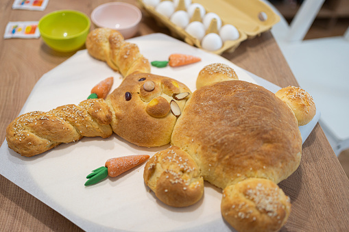 Baked handmade bread in a shape of an Easter Bunny on a plate, eggs, and color dye on the kitchen counter\nEaster season
