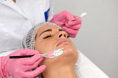 Beautician uses brush to apply white mask made of kaolin clay on half of woman's face. Close up of young woman in white headband lying on soft towel in beauty salon. Concept of cosmetic care for face