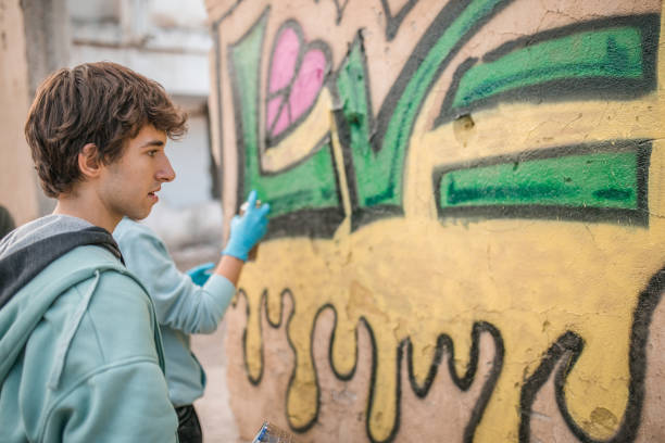 Cute young woman graffiti artist dressed in casual clothes is painting the mural in street art style on the large sheet. She is working paintbrush. Studio shooting