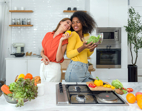 Two women stand in a kitchen holding various fruits and vegetables, smiling.