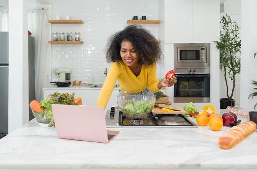 A young woman stands in a kitchen with a laptop and vegetables, likely cooking or looking up recipes.