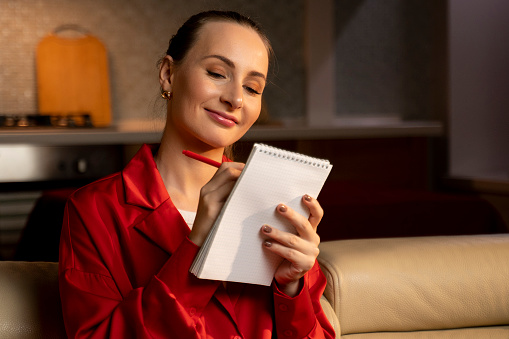 A woman dressed in a vibrant red blaser thoughtfully touched her face with a pen while holding a notepad, looking away in a warmly lit indoor setting.